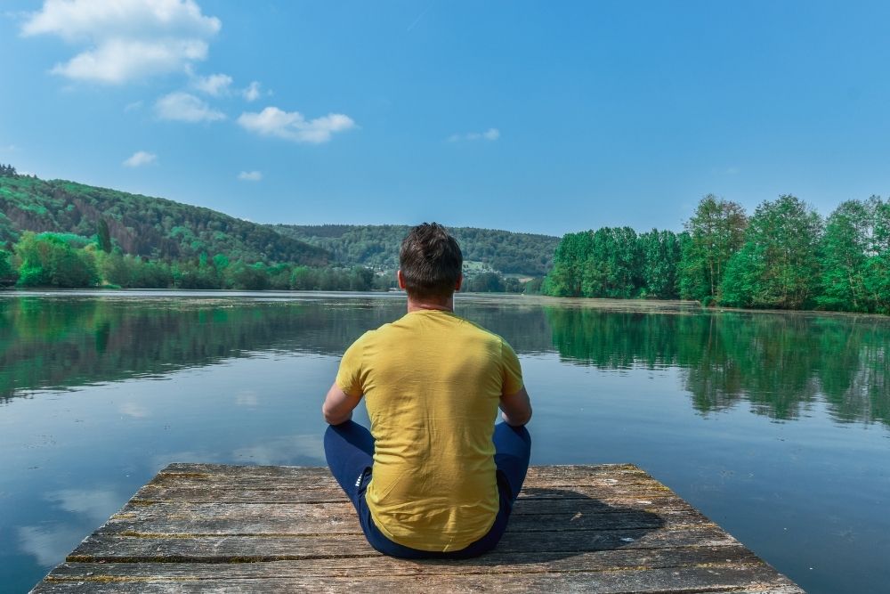 A man sitting calmly by a lake, practicing mindfulness for sustainable fitness and body awareness.