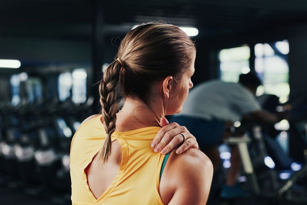A woman in a yellow tank top massages her aching shoulder in a gym