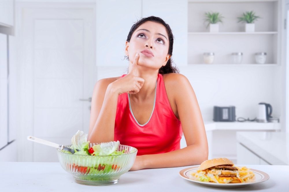A thoughtful person in a kitchen with a salad and a burger, representing the decision-making process in daily nutrition.