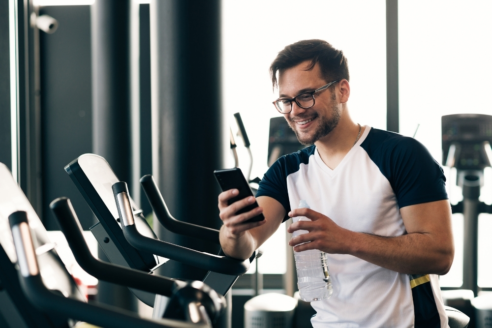 An athlete at the gym using a smartphone to check workout stats and connect with their coach, representing a hybrid approach to fitness.