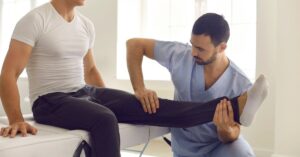 A male physical therapist in blue scrubs performs passive range of motion therapy on a male patient's leg.