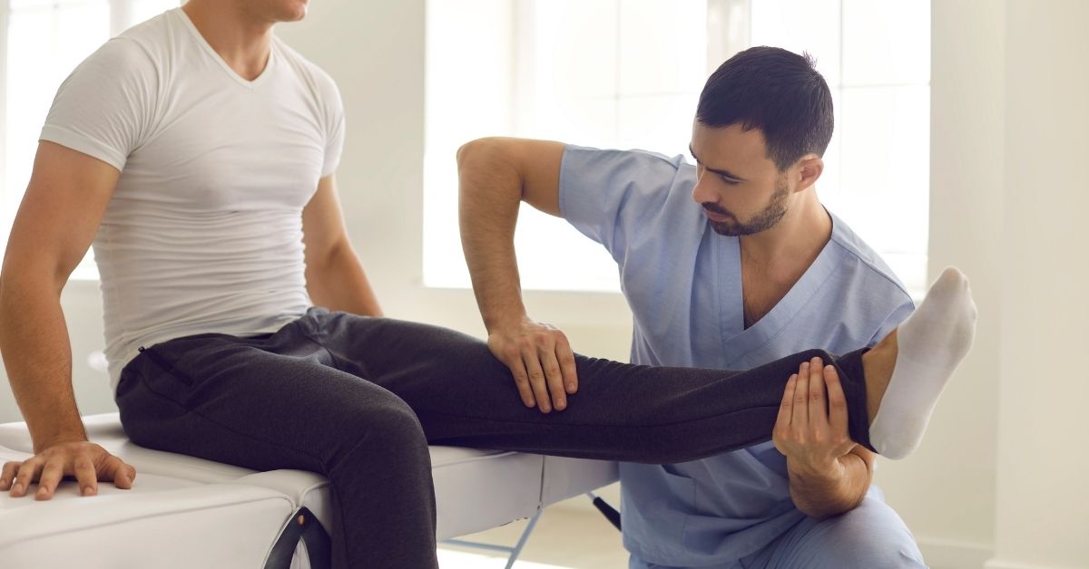 A male physical therapist in blue scrubs performs passive range of motion therapy on a male patient's leg.