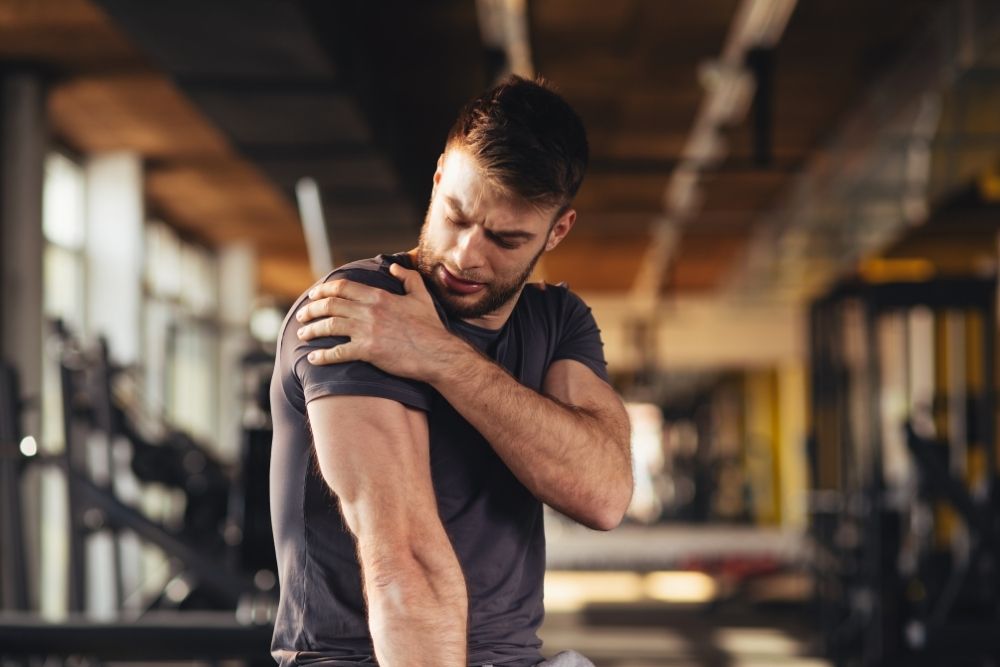 A man in a gym holds his left shoulder in visible pain, indicative of a rotator cuff injury or strain.