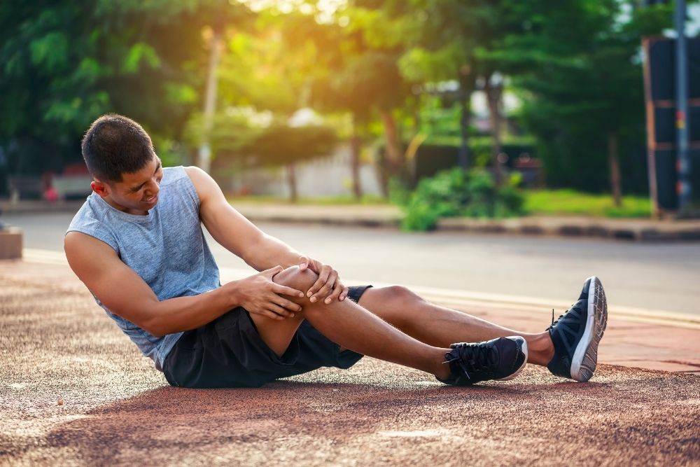 A male runner sits on an outdoor track, clutching his injured knee, suggesting a sports-related injury.