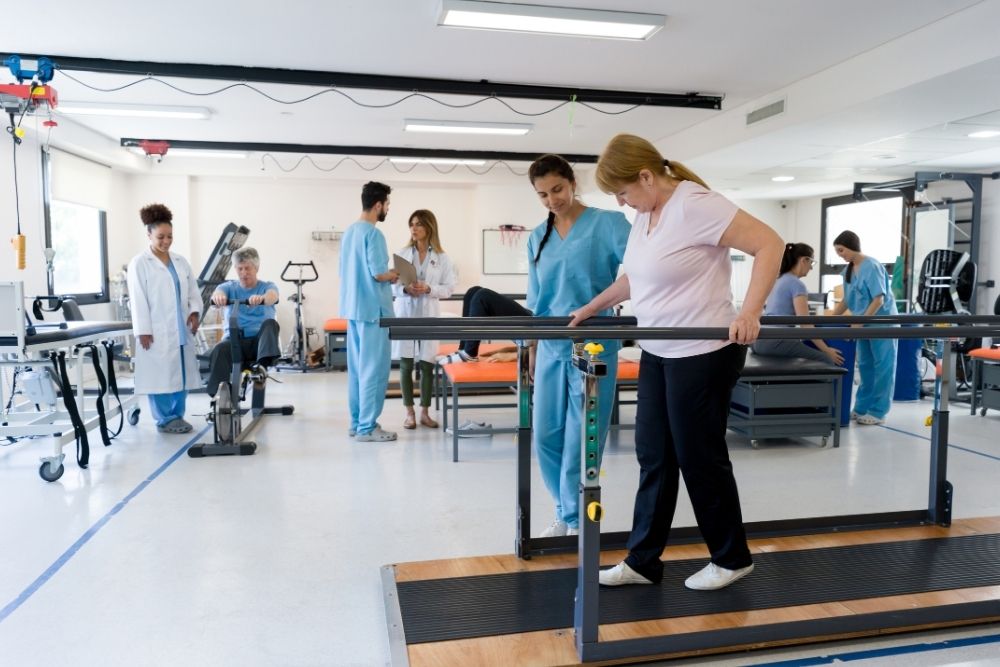 A female patient in a physical therapy clinic uses parallel bars to practice walking with the assistance of a female therapist, focusing on lower body injury recovery.