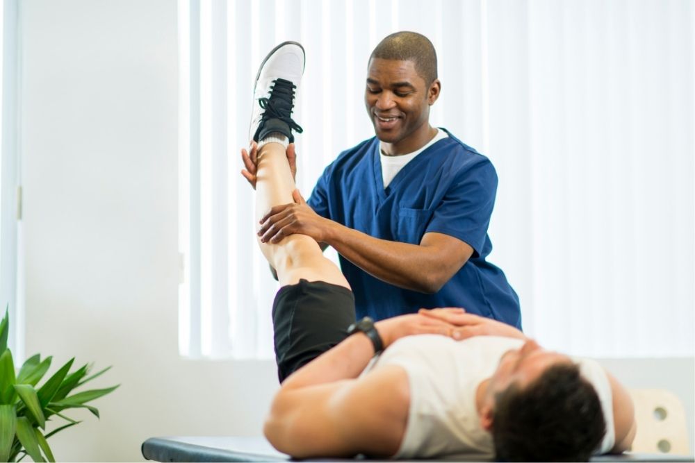 A male physical therapist in blue scrubs stretches the hamstring of a male patient lying on a treatment table, aiding in injury recovery.