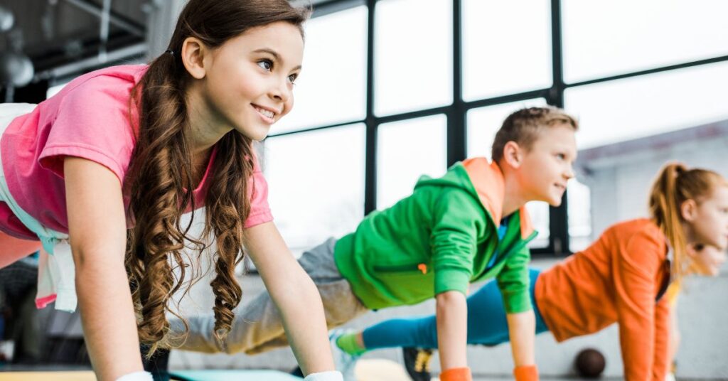 A group of children smiling and doing core planks together as part of a structured kids and teens fitness class.