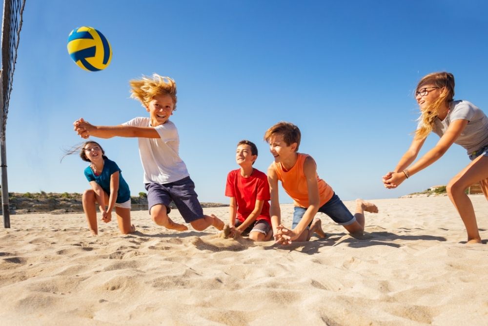 A group of kids and teens playing volleyball together on the beach, showcasing active hobbies in kids and teens fitness.